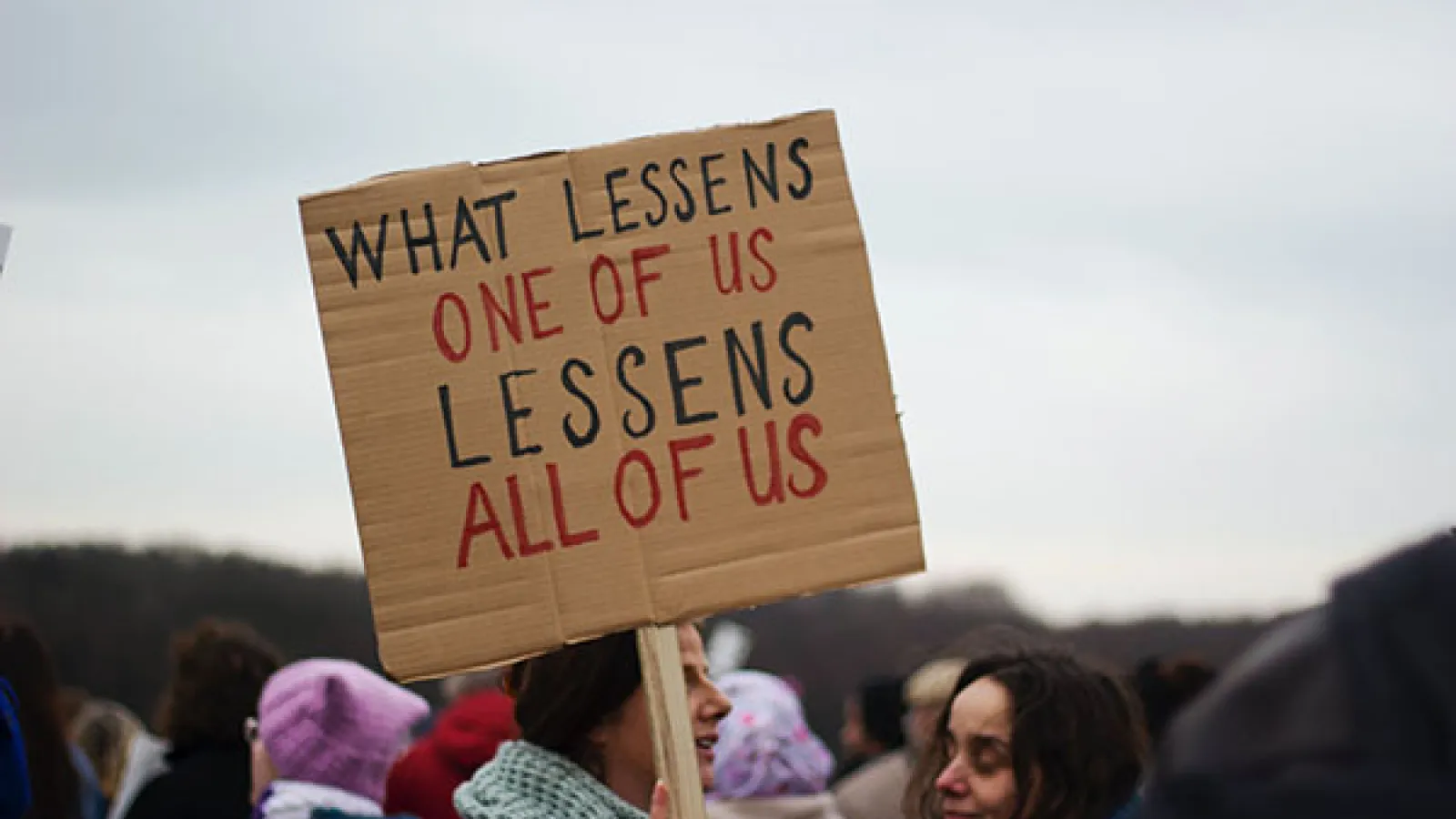 Women with protest sign that reads "What lessens one of us lessens all of us"