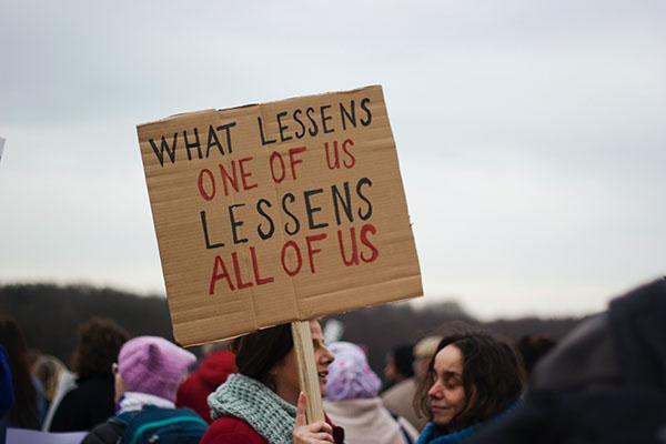 Women with protest sign that reads "What lessens one of us lessens all of us"